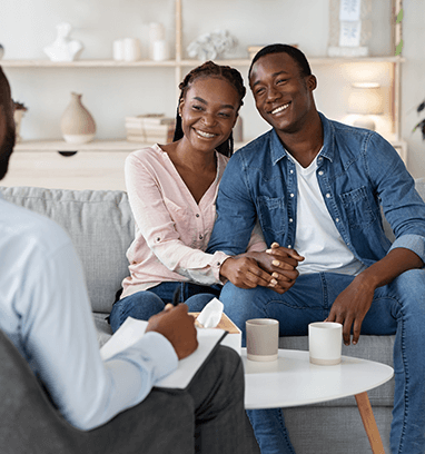 Couple smiling and holding hands across from a therapist in a bright living room