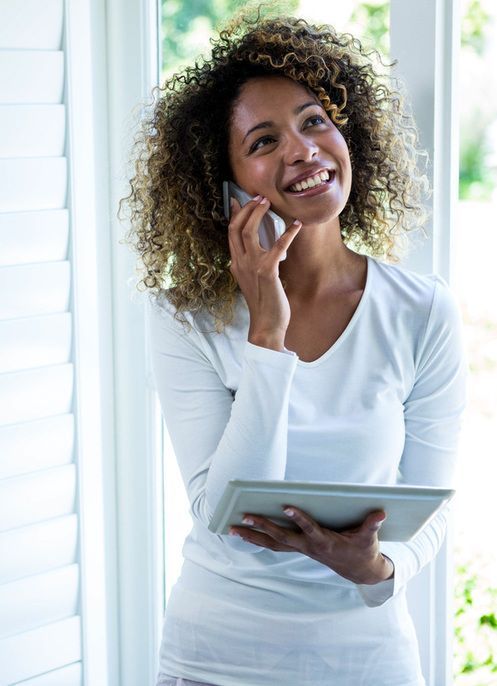 Smiling person on a phone, holding a tablet in a bright room near a window.