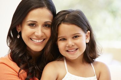 Smiling woman and young girl posing together indoors, both in light-colored tops.