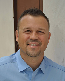 Smiling man in a light blue button-up shirt standing indoors near a doorway