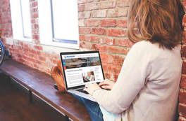 Person using a laptop on a wooden bench by a brick wall and window in a bright room