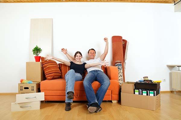 Two people sitting on an orange couch in a bright living room, smiling with arms raised among moving boxes
