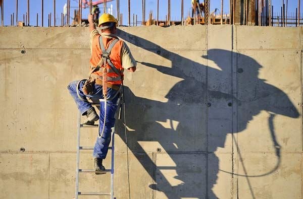 Construction worker on a ladder beside a concrete wall, casting a large shadow in sunlight.