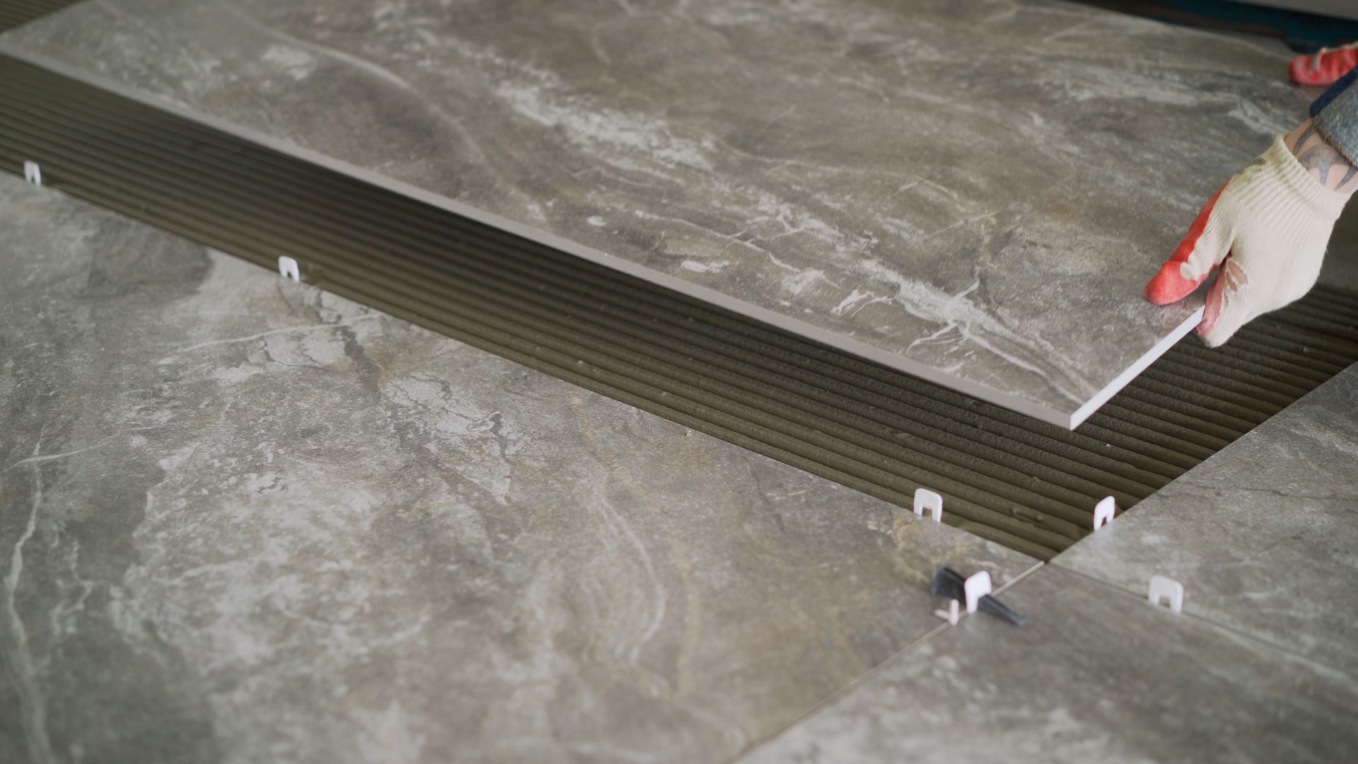 A gloved hand placing a gray tile onto a surface with tile leveling clips