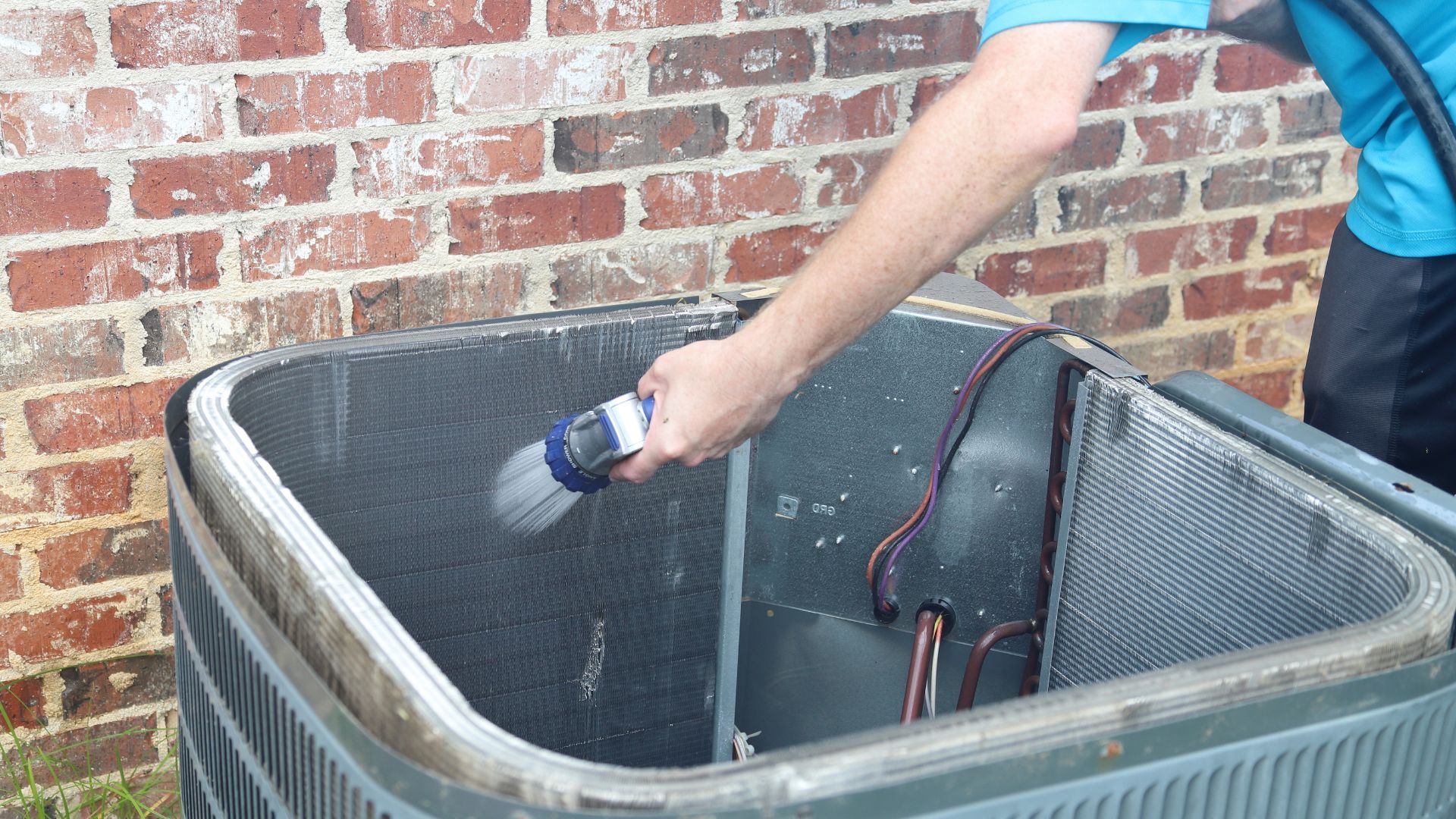 Man cleaning an air conditioner's coils with a brush, near a brick wall.