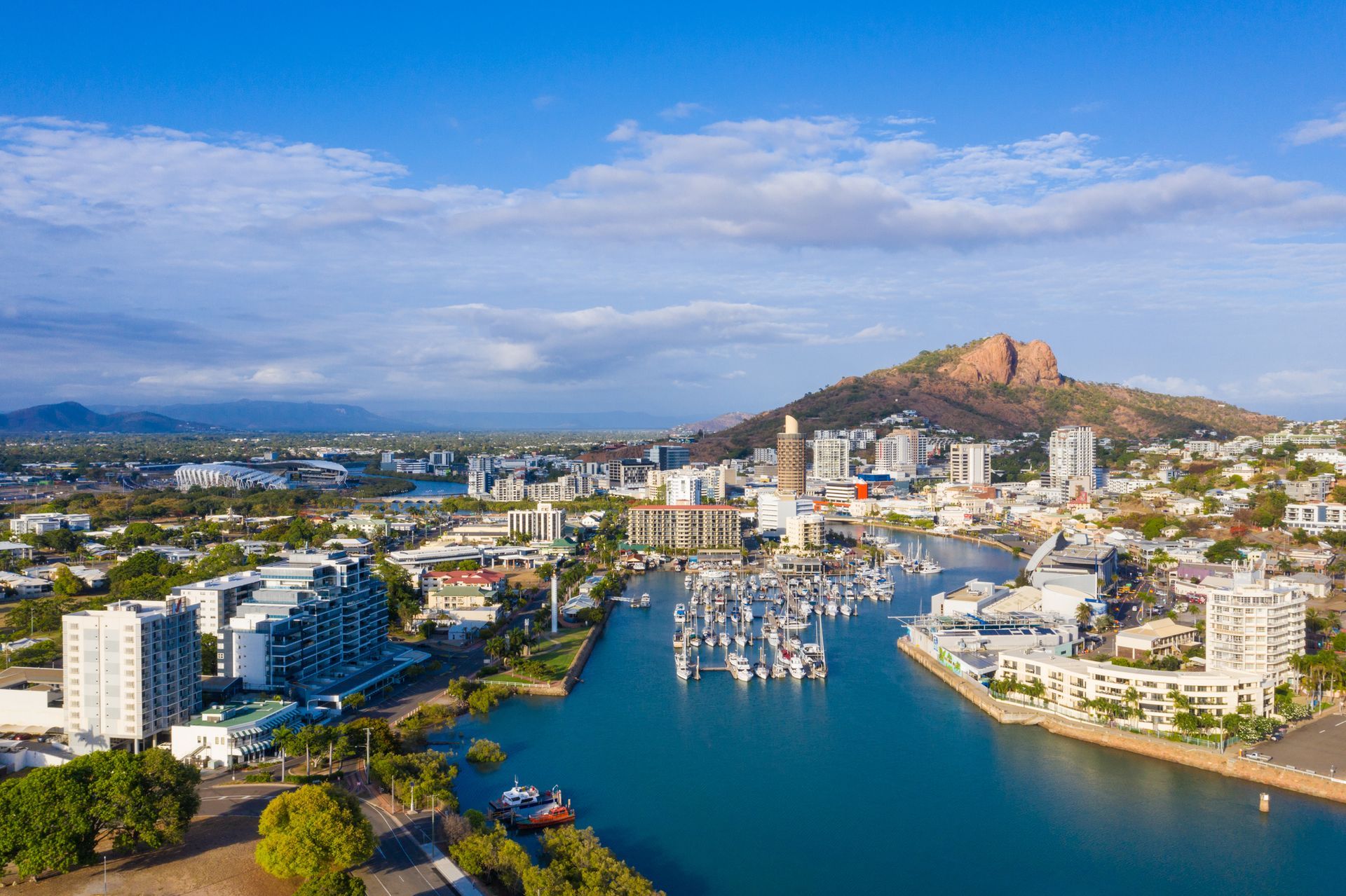 Aerial view of a coastal city with boats in a harbor, surrounded by buildings and a mountain under a blue sky.