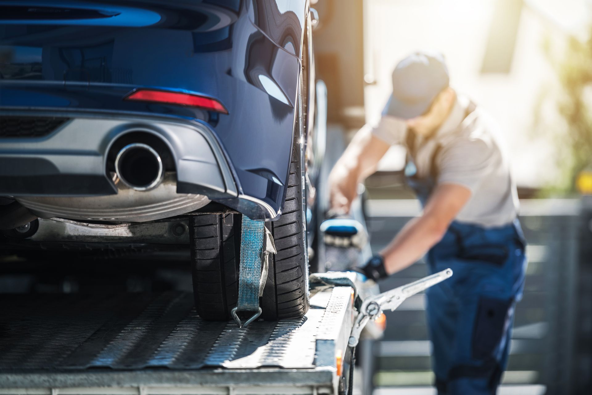 A tow truck driver securing a blue car onto a flatbed truck. Sunny outdoors.