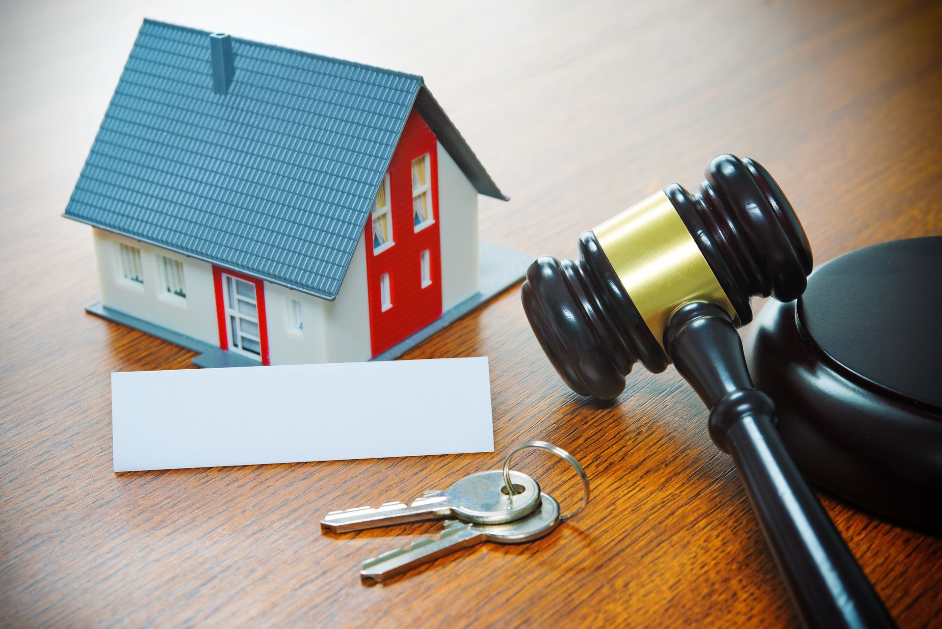 Miniature house, gavel, keys, and blank sign on wooden table, possibly representing a real estate auction.