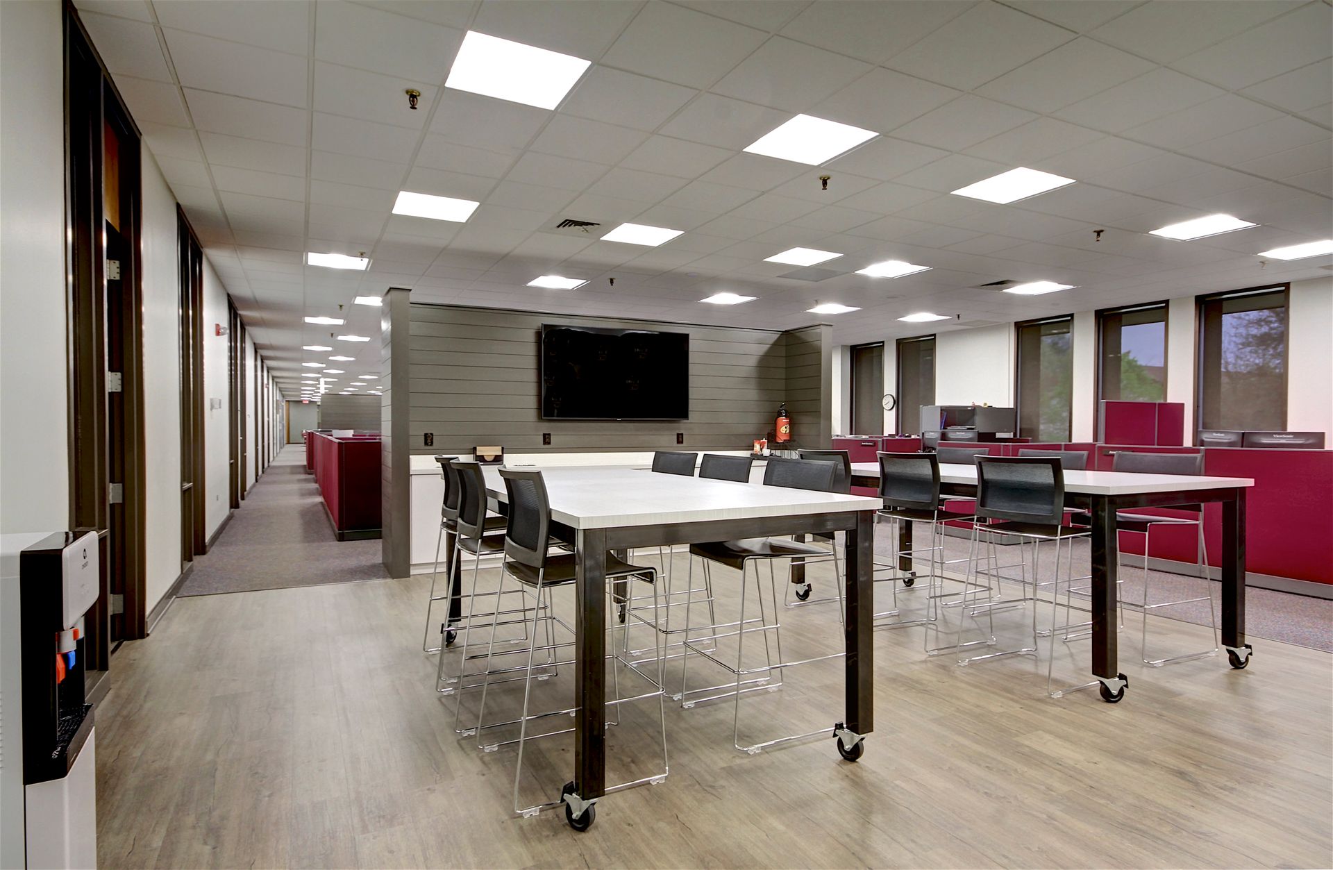 Modern open office with long tables, chairs, bright ceiling lights, and windows along the wall.