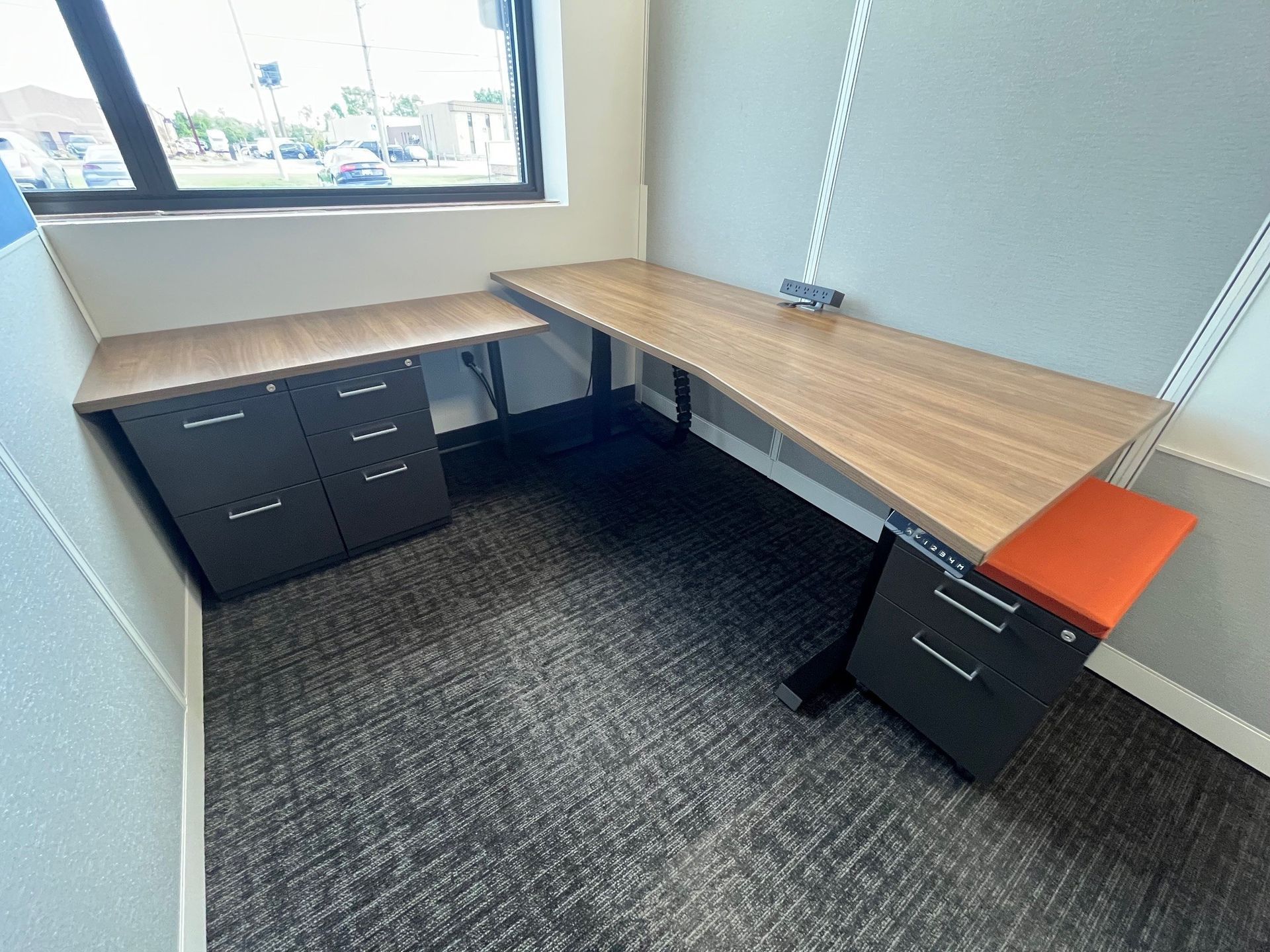 Small office corner with L-shaped wood desks, black drawers, orange chair, and a window.