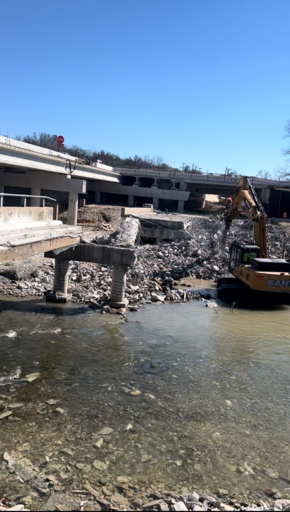 Demolition excavator clearing rubble beside a shallow river under a partly destroyed concrete building.
