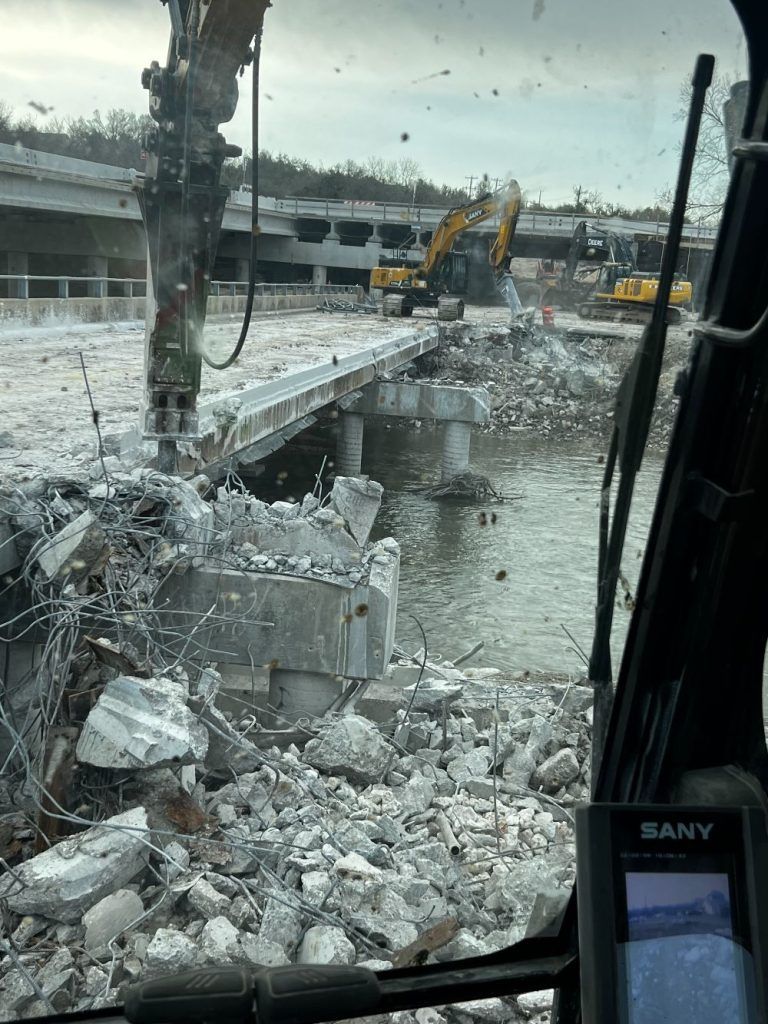 Demolition site with rubble, broken concrete, and excavators beside a damaged bridge over water