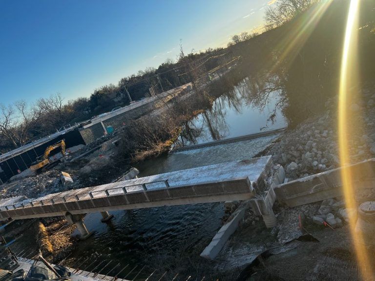 Collapsed bridge over a partially frozen river at sunset, with debris and a strong sun glare.