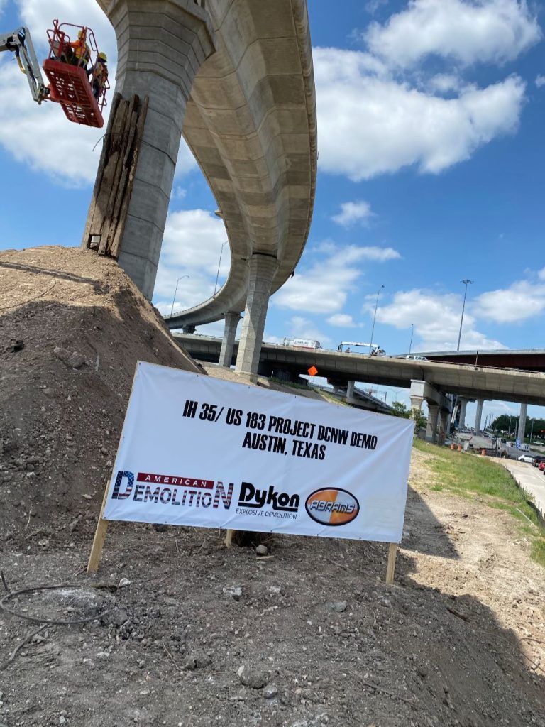 Banner on a dirt embankment beneath a highway overpass under a blue sky with clouds.