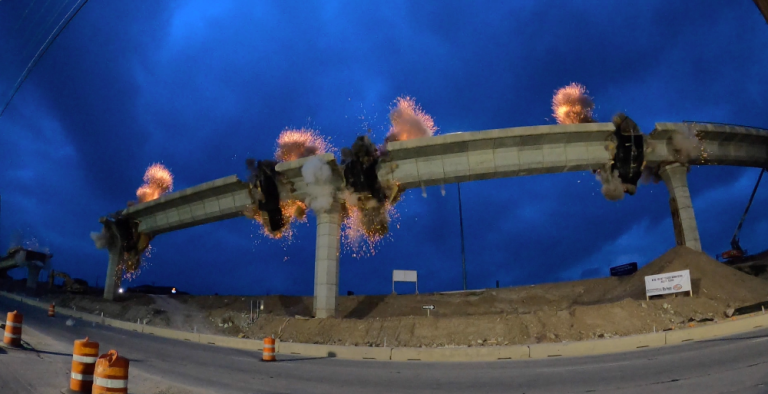 Damaged elevated roadway collapsing in flames at night, with orange construction barrels below.