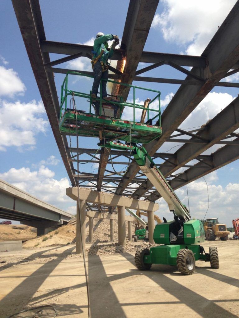 Construction workers on a green boom lift installing steel beams under a partially built structure.