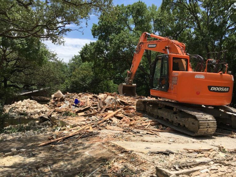 Orange excavator clearing debris in a wooded area under trees