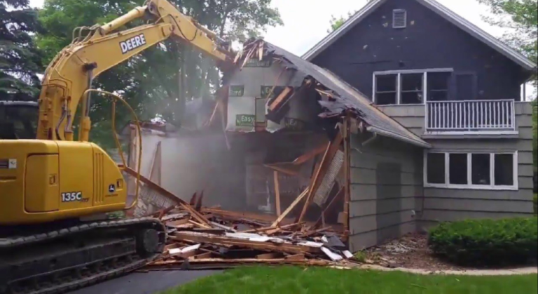 Yellow excavator demolishing a house section, with debris and dust in a suburban yard