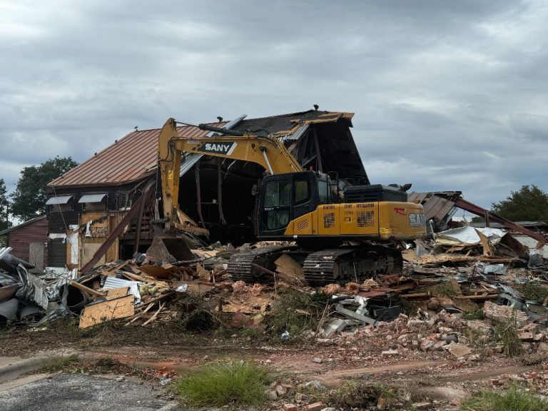 Yellow excavator demolishing a collapsed brick house amid rubble and debris under a cloudy sky