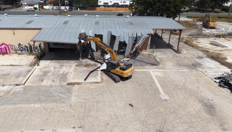 Excavator demolishing a low building with debris scattered across a concrete lot
