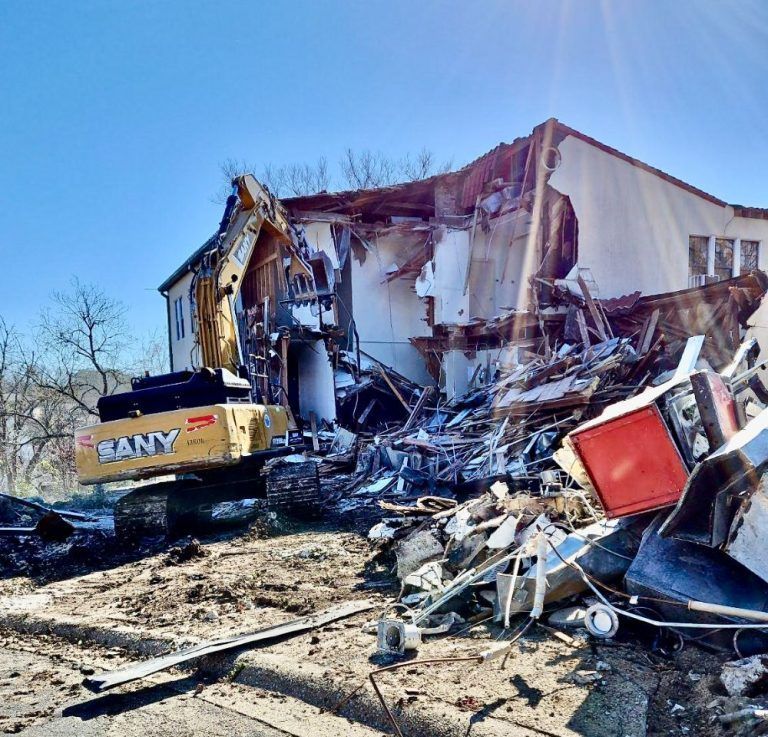 Excavator amid rubble beside a partially demolished building under a clear blue sky