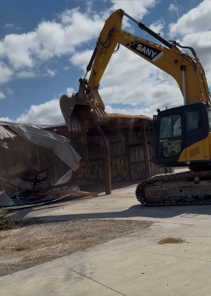 Yellow excavator tearing down a corrugated metal building under a cloudy sky