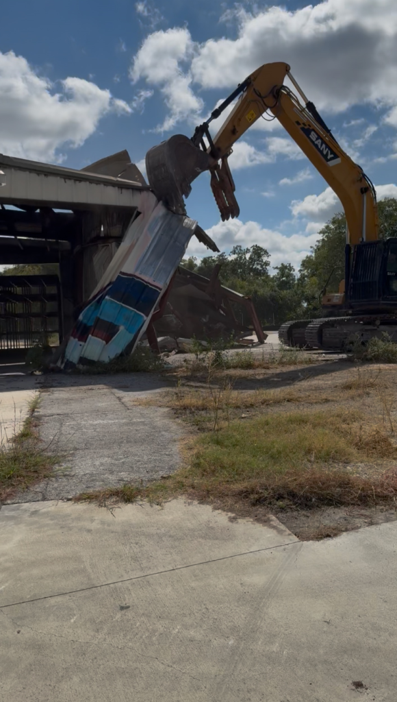 Excavator lifting a blue bus at a demolition site under a partly cloudy sky