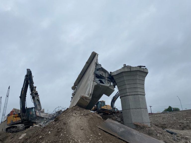 Collapsed bridge deck with construction machinery and debris under a cloudy sky