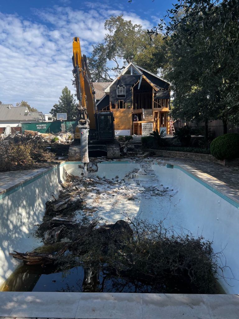Excavator clearing debris from an empty backyard pool beside a damaged house under repair