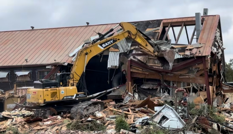 Yellow excavator demolishing a damaged building with debris scattered in front.