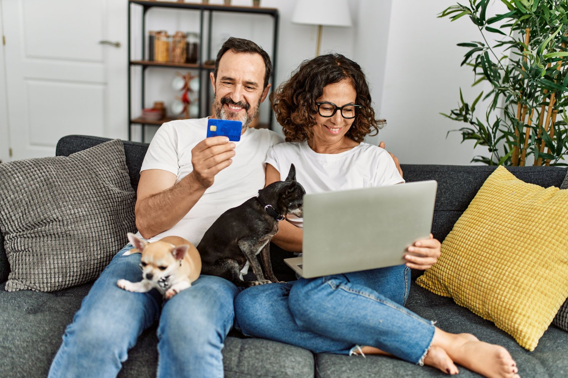 Couple on a couch using a laptop and phone with two small dogs curled up beside them