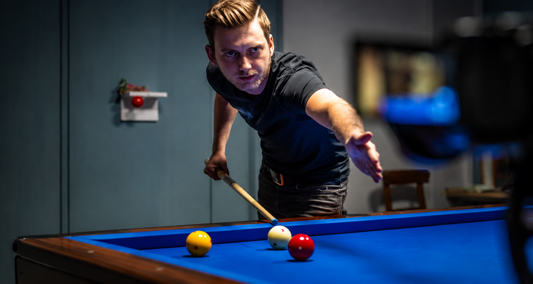 Een man in een zwart shirt richt met een biljartkeu op biljartballen op een blauwe tafel.