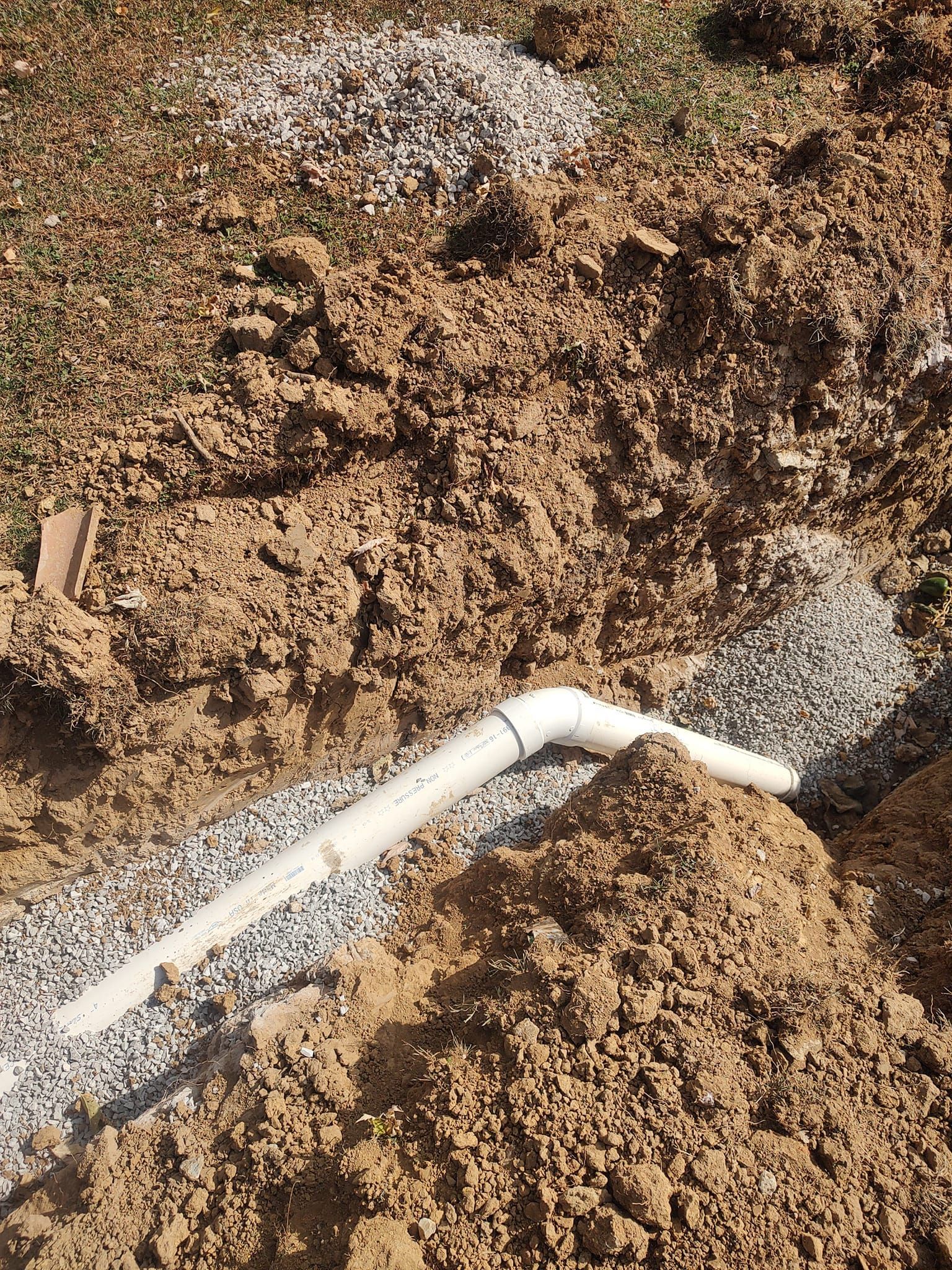 White PVC pipe in a trench filled with gravel, surrounded by brown soil and more gravel on the surface.