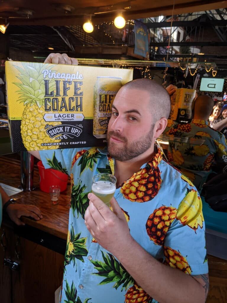 Person in pineapple print shirt with case of Life Coach Lager with Pineapple beer on their shoulder