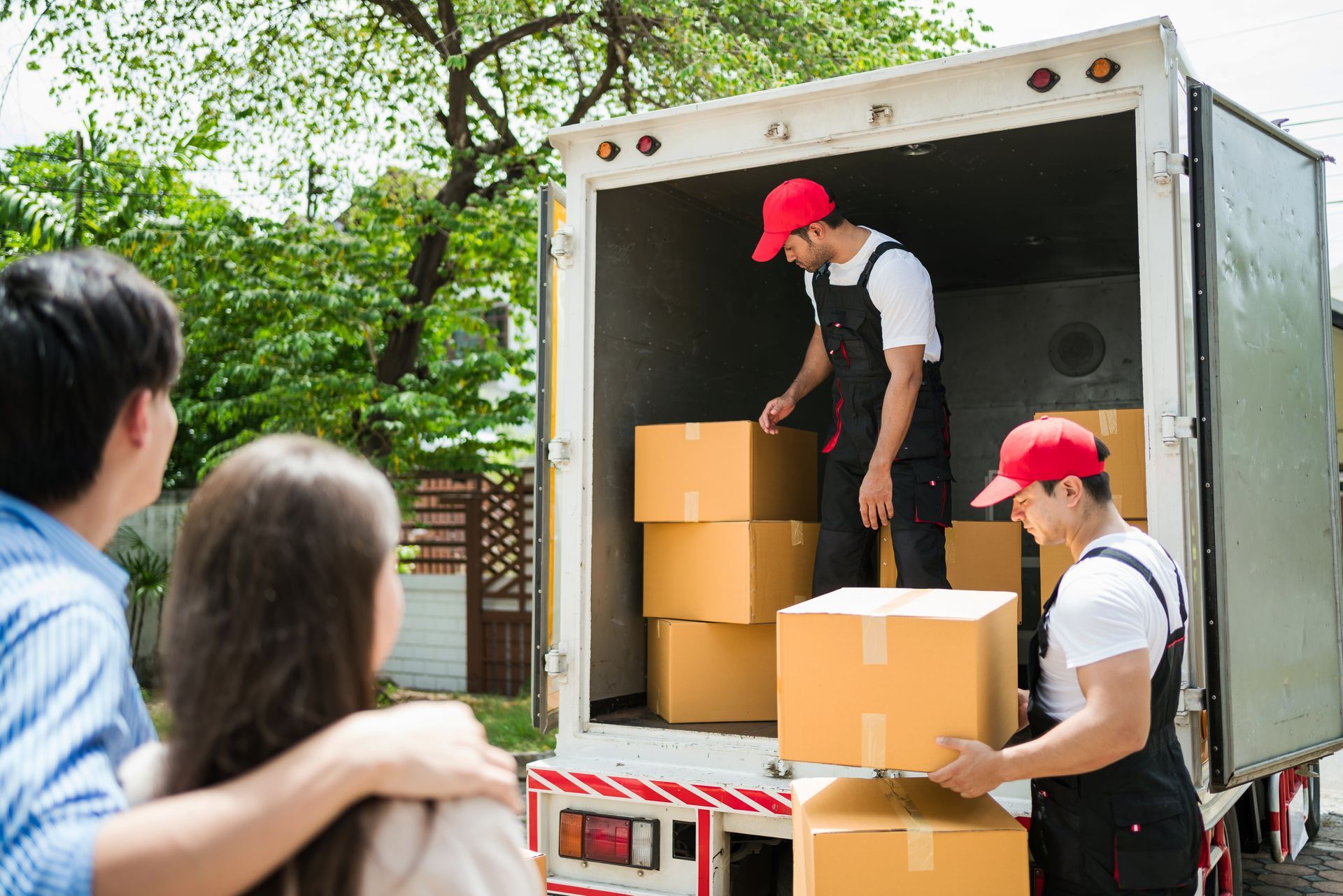 Movers loading cardboard boxes into a truck while two people watch from the sidewalk.