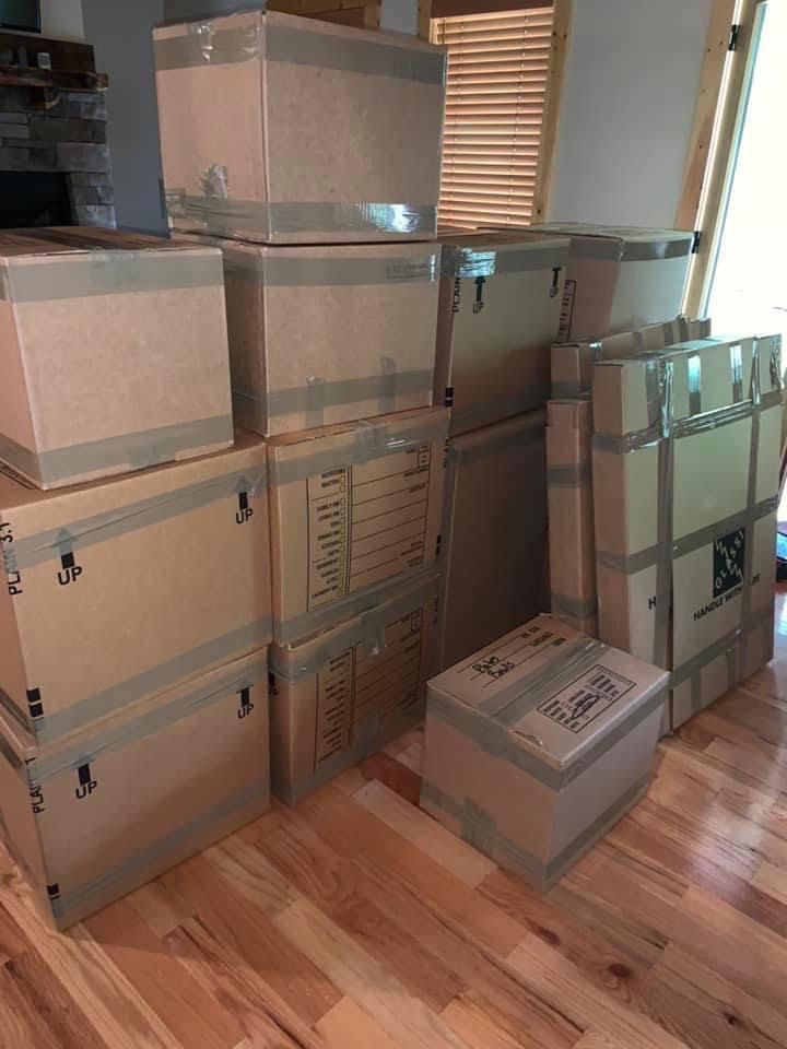 Delivery men loading cardboard boxes - Deep Gap, NC - Appalachian Moving & Storage Stacked cardboard boxes and containers in a cluttered room with wooden floors and blinds.