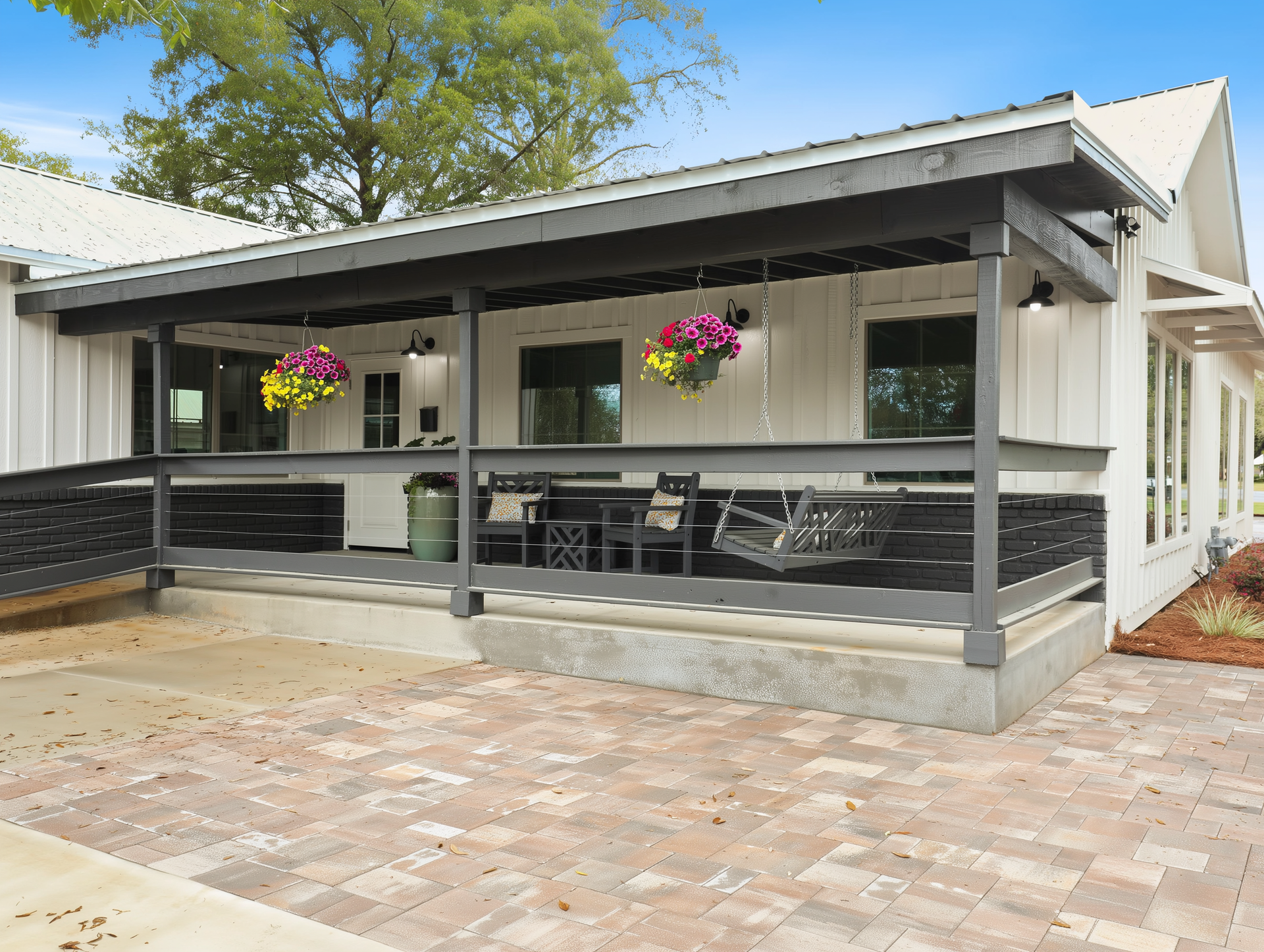 Covered patio with gray railings, hanging flower baskets, and outdoor seating beside a paved driveway
