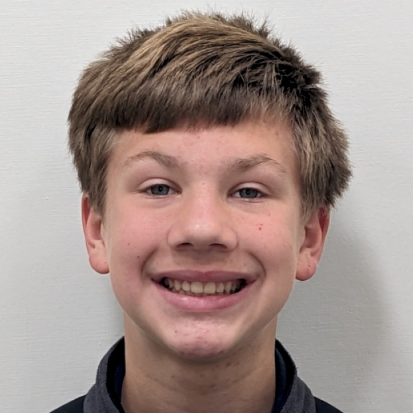A smiling person with light brown hair and blue eyes, wearing a black hooded sweatshirt against a plain gray background.