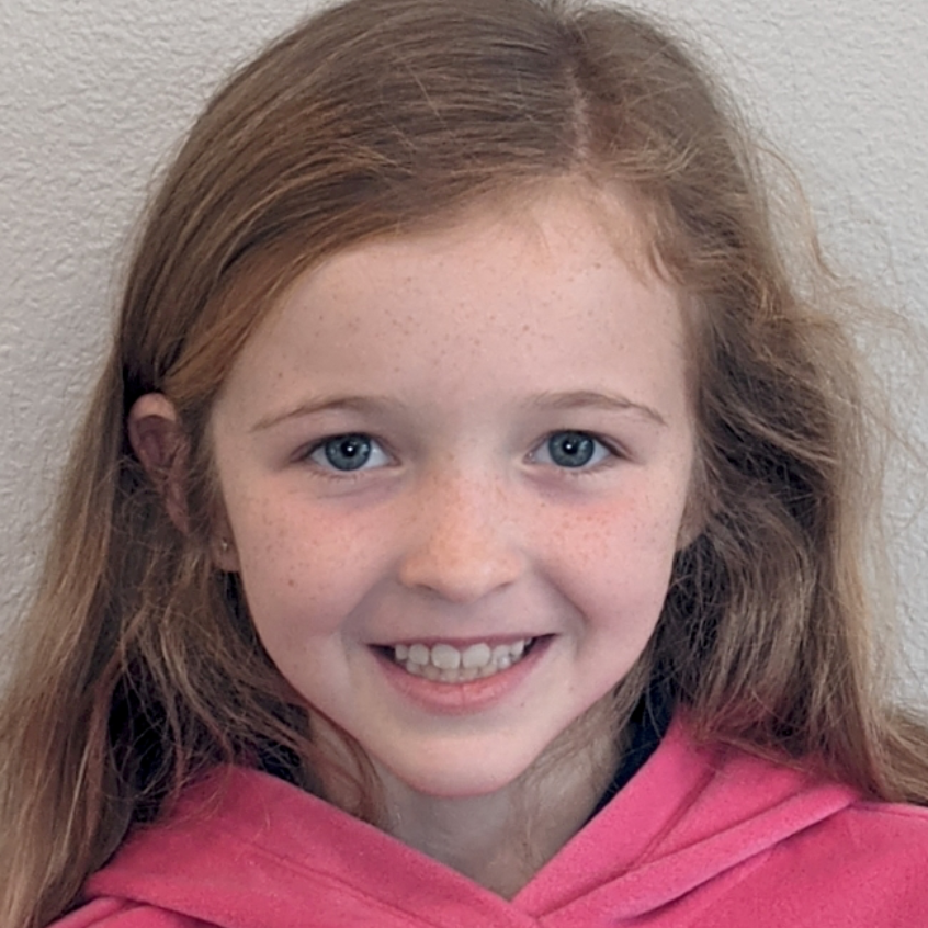 A person with long blonde hair and light skin smiling at the camera, wearing a pink hooded sweatshirt against a plain wall.