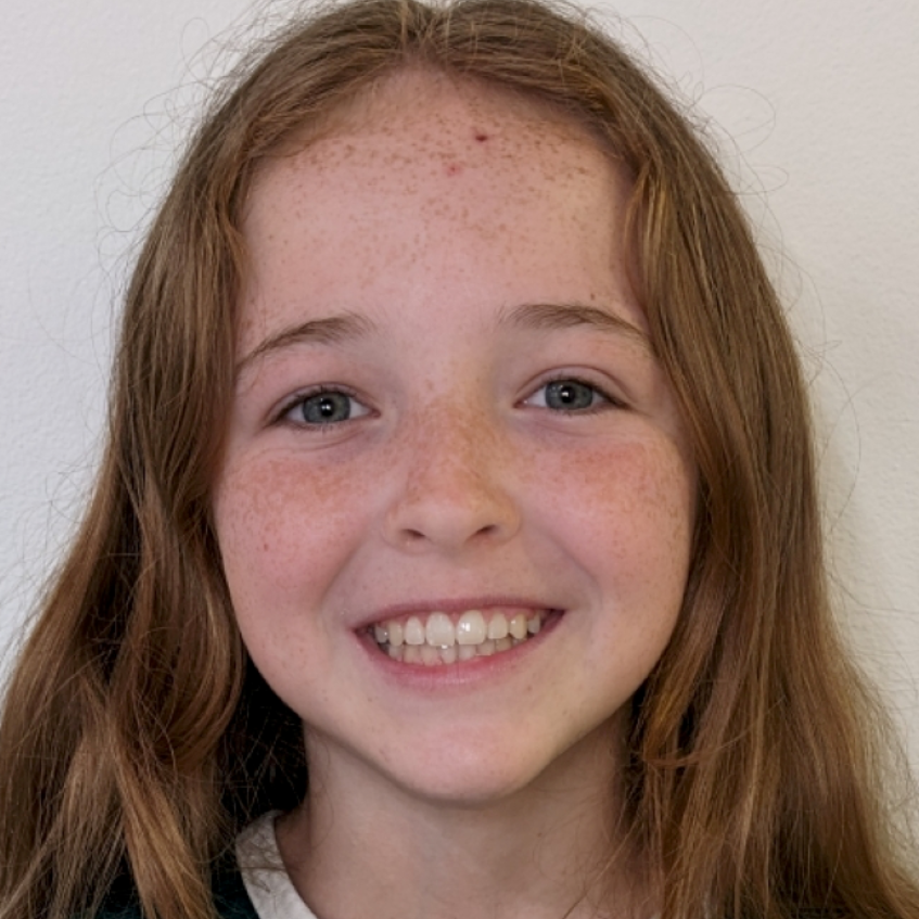 A smiling child with long reddish-brown hair and light skin with freckles faces forward against a plain white background.