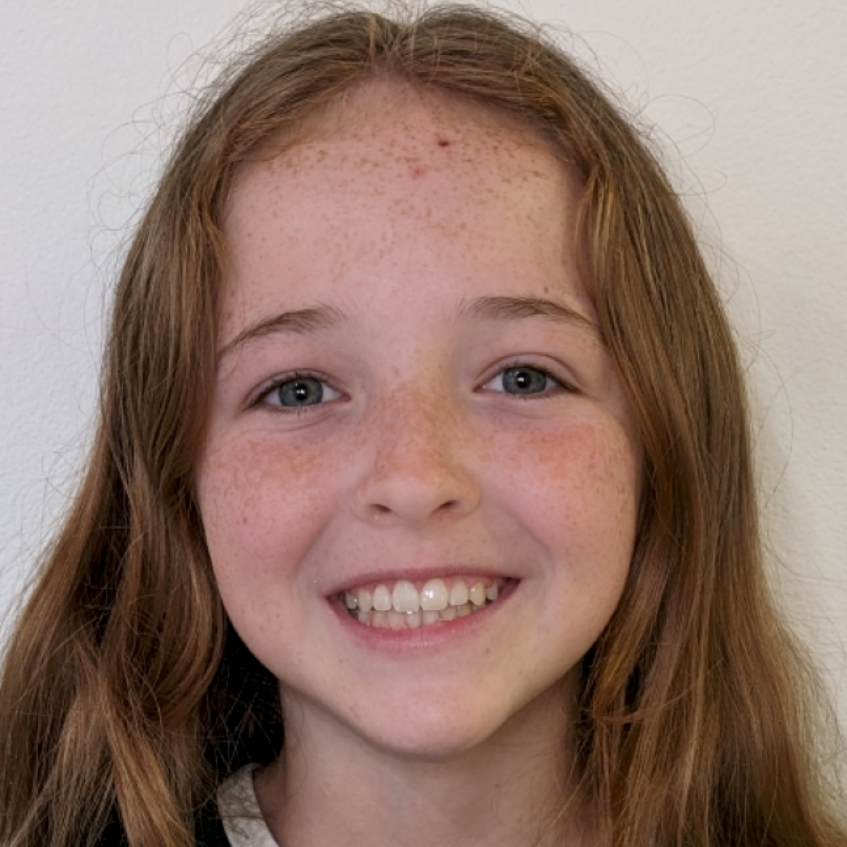A headshot of a person with long, light-brown hair and freckles smiling broadly against a plain white background.
