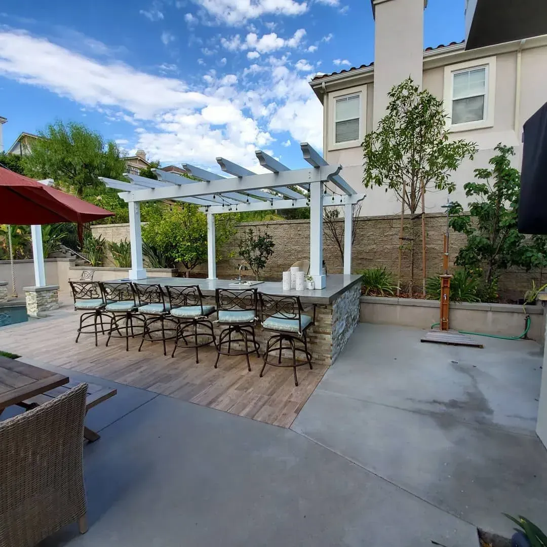 Outdoor bar with seating under a pergola, adjacent to a patio and a swimming pool. Blue sky.