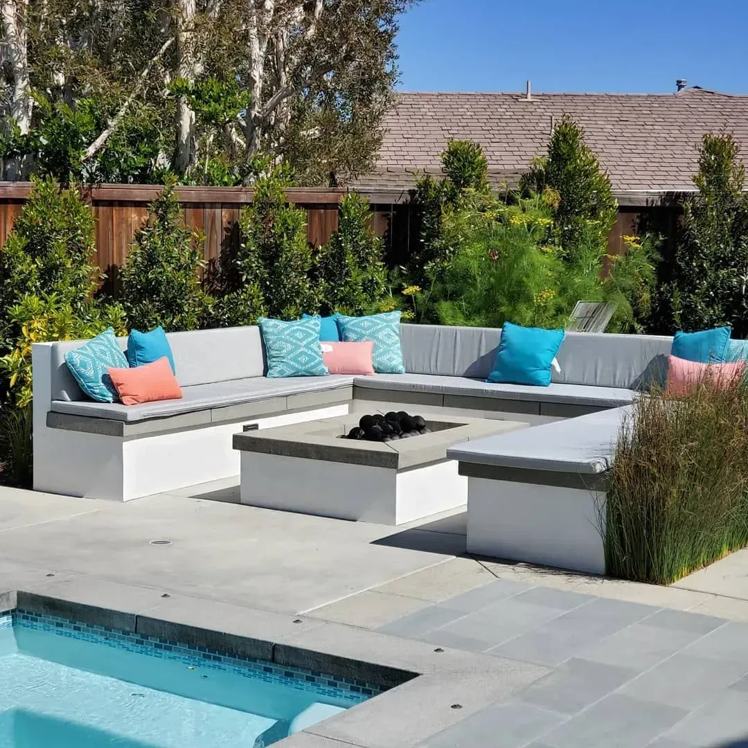 L-shaped concrete outdoor seating area with cushions around a fire pit and a pool in a backyard.