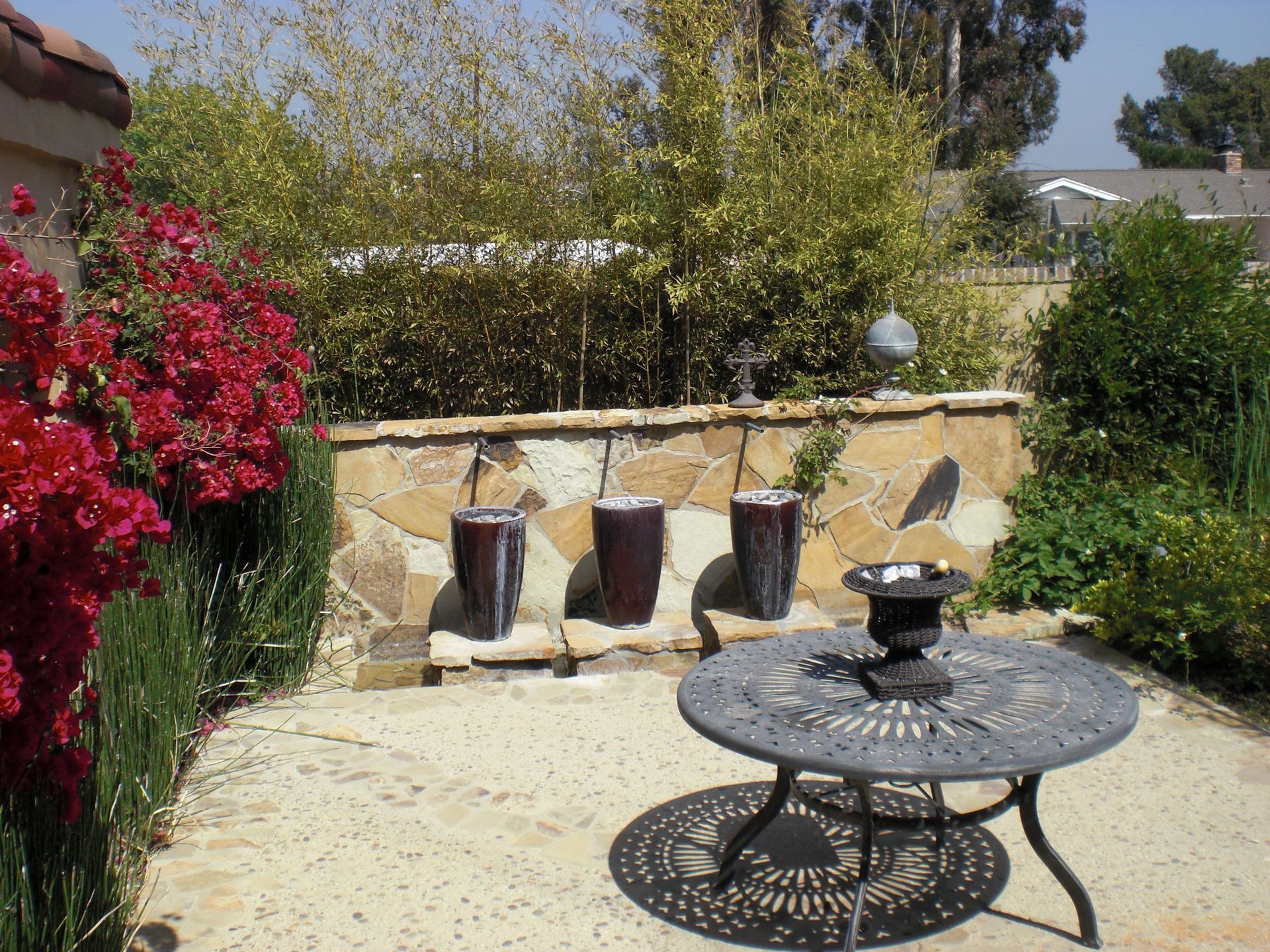 Patio with red flowering bushes, stone wall fountain, metal table, and plants.