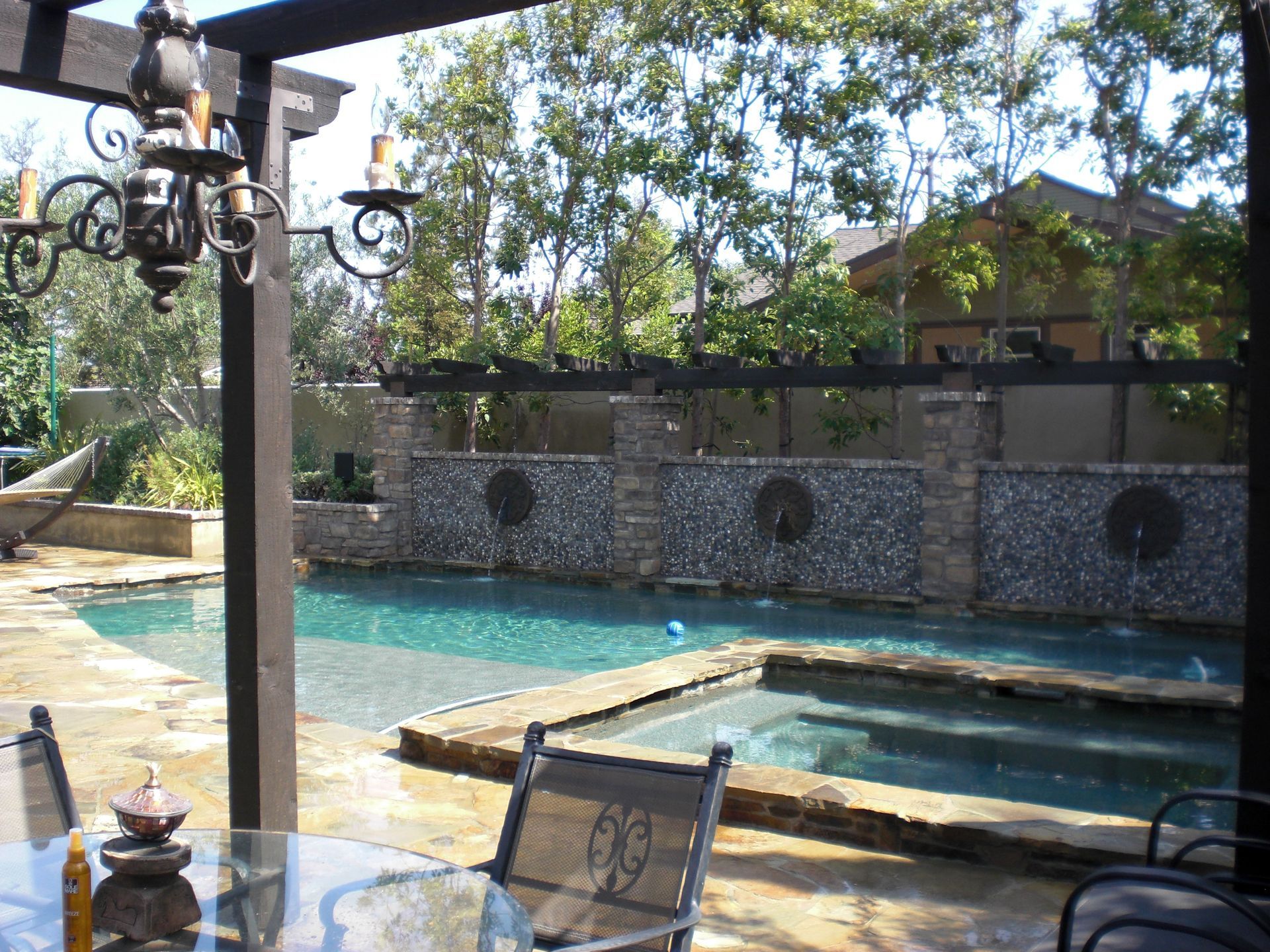 Pool and spa with stone wall backdrop, under a wooden pergola, daytime.