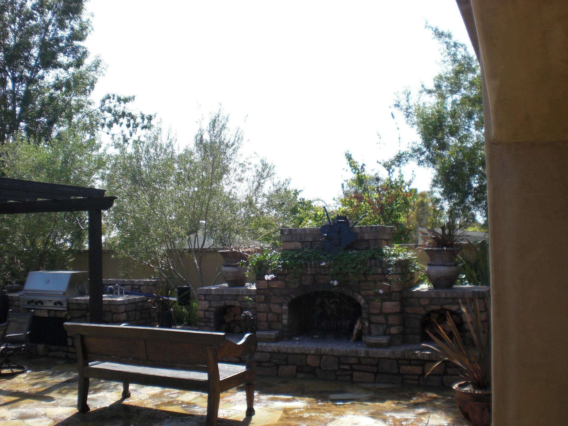 Outdoor patio with a stone fireplace, grill, seating, and trees.