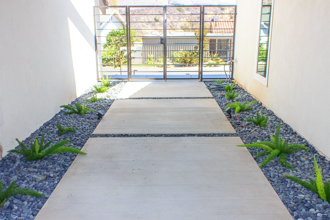 Concrete walkway with flanking beds of dark gravel and plants, leading to a metal gate.