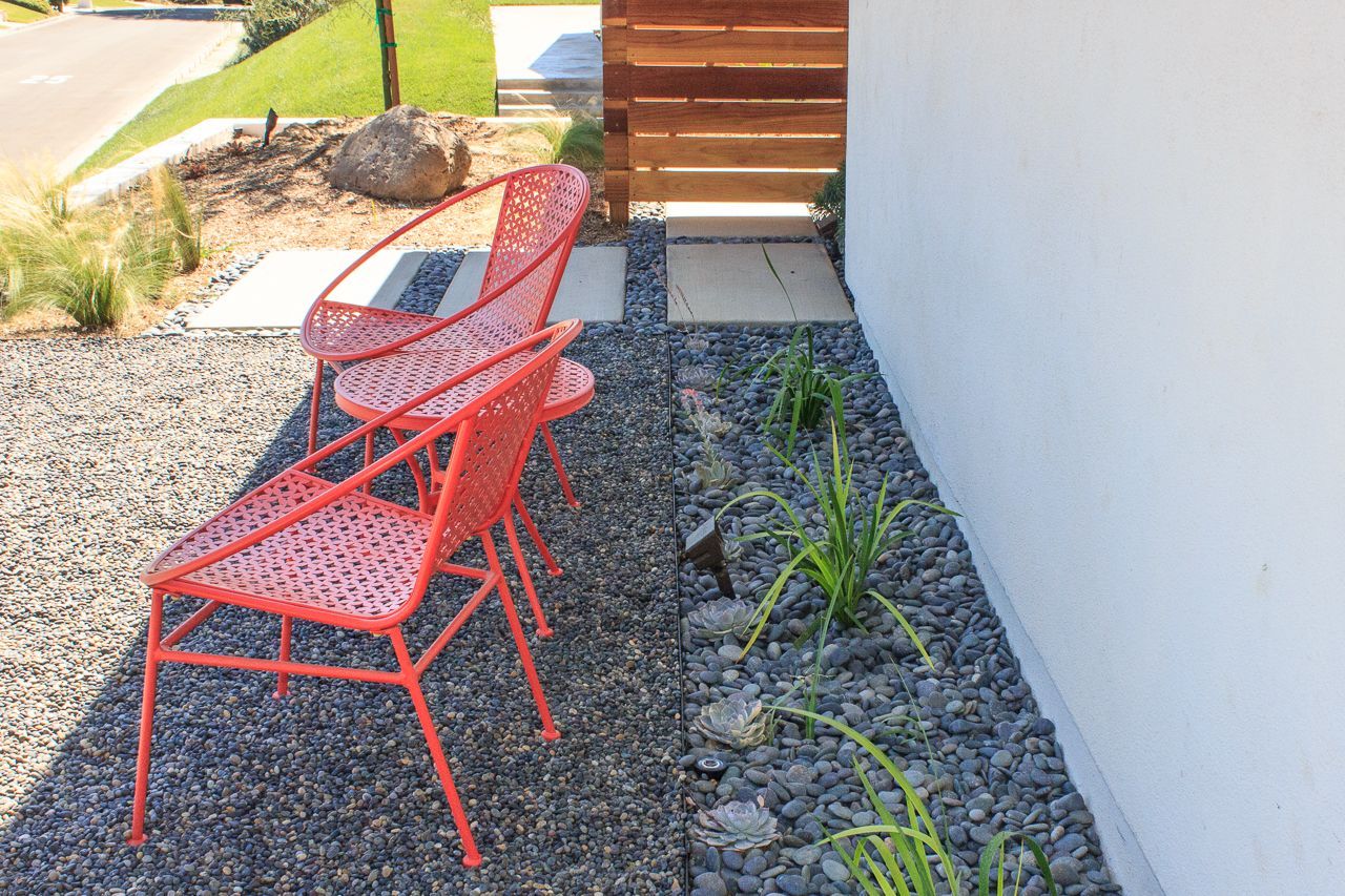 Two red metal chairs sit on gravel beside a white wall and wooden fence.