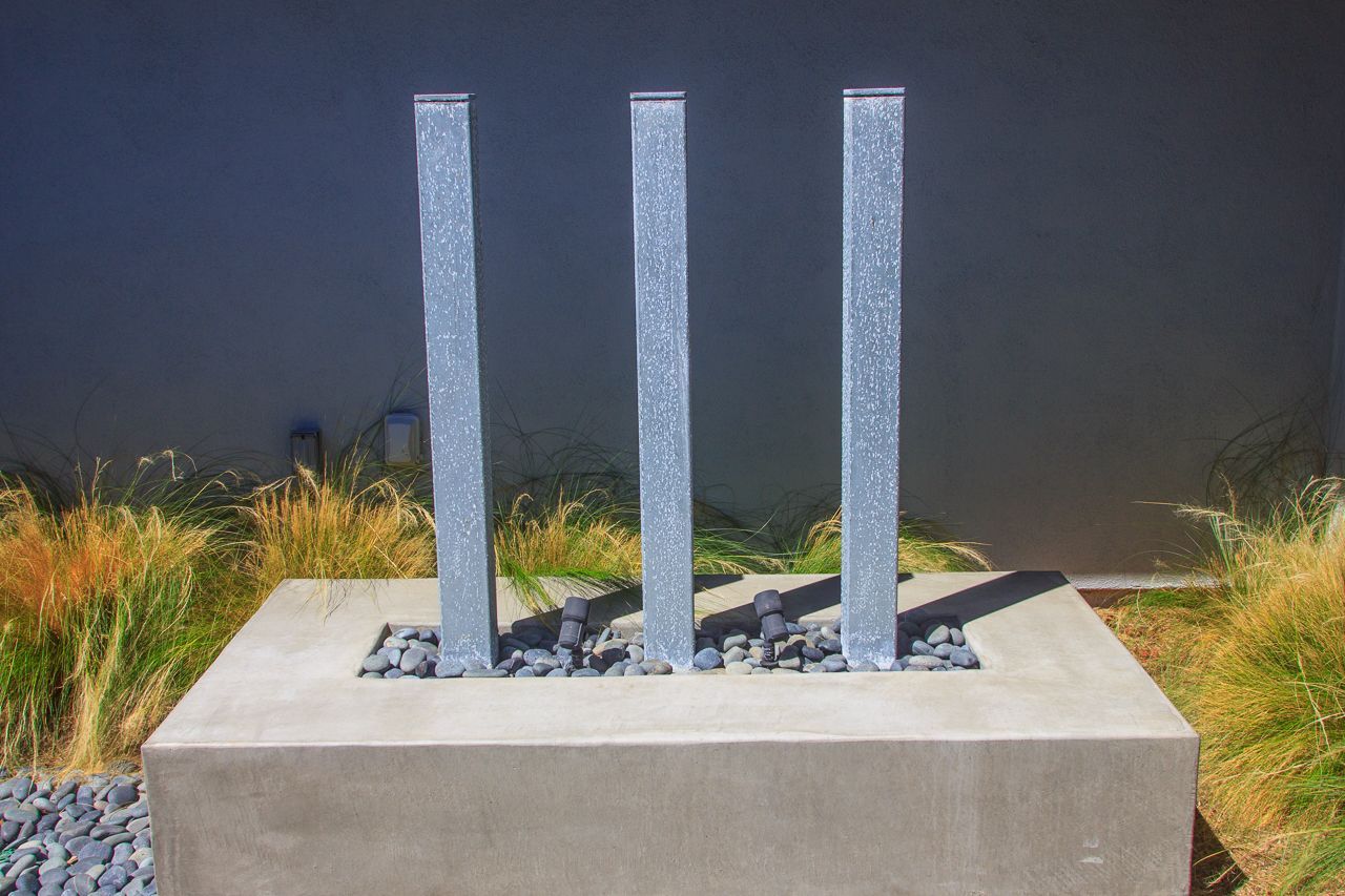 Three water columns in a square concrete basin, surrounded by rocks and ornamental grass.
