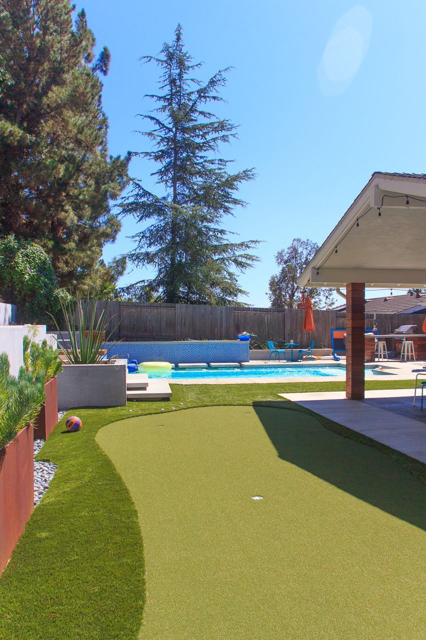 Backyard with putting green, pool, and patio under a bright blue sky.